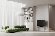 A modern living room with a minimalist design features a green modular sofa set against a backdrop of sheer white curtains. A large white dome light fixture hangs above. To the side, a tall bookshelf filled with various books is integrated into the wall. A sleek, black flat-screen TV rests on a stand with slender, wooden legs, adjacent to a small staircase.