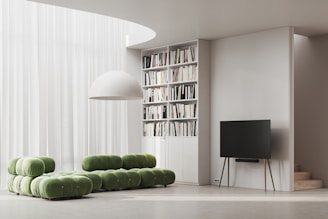 A modern living room with a minimalist design features a green modular sofa set against a backdrop of sheer white curtains. A large white dome light fixture hangs above. To the side, a tall bookshelf filled with various books is integrated into the wall. A sleek, black flat-screen TV rests on a stand with slender, wooden legs, adjacent to a small staircase.