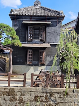 A traditional Japanese-style building with dark wooden elements and a tiled roof, surrounded by a wooden fence. Stacked logs are aligned neatly in front of the building. A meticulously trimmed pine tree and another small tree with slender leaves add greenery to the scene. The sky is bright and clear with a few small clouds.