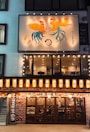 A Japanese izakaya with illuminated signage and lanterns. The upper portion features a decorative panel with two stylized roosters and skewers. The entrance is adorned with traditional Japanese lanterns and displays menus on either side. Inside, patrons are visible through wooden-framed glass doors.