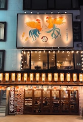 A Japanese izakaya with illuminated signage and lanterns. The upper portion features a decorative panel with two stylized roosters and skewers. The entrance is adorned with traditional Japanese lanterns and displays menus on either side. Inside, patrons are visible through wooden-framed glass doors.