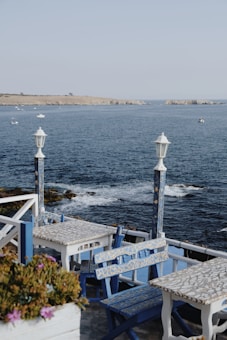 A coastal scene featuring a charming outdoor caf&eacute; with blue and white mosaic tables and chairs. The caf&eacute; overlooks a serene body of water with gentle waves lapping the shore. Several small boats are visible in the distance, and a rocky coastline stretches out under a clear sky. Two white lampposts frame the view, and a planter with pink flowers adds a splash of color to the foreground.