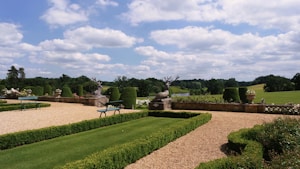 A beautifully manicured garden with lush green grass, neatly trimmed hedges, and gravel paths. Stone sculptures of deer with antlers sit atop a stone wall. Benches are positioned on either side for seating, surrounded by vibrant flowers and potted plants. The landscape extends to include rolling hills and a blue sky filled with fluffy clouds.