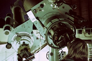 A large, complex telescope with detailed mechanical parts and optics is situated in a dimly lit room. A person with long hair stands close to the telescope, suggesting observation or adjustment. The setting appears industrial or scientific, with visible gears and controls.