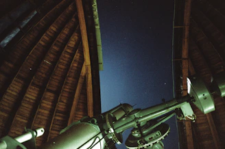 A large telescope is positioned inside an observatory with an open wooden roof, looking out towards a clear night sky with faint stars visible.