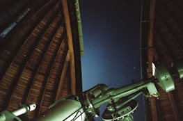 A large telescope is positioned inside an observatory with an open wooden roof, looking out towards a clear night sky with faint stars visible.
