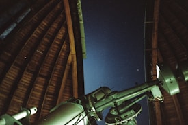 A large telescope is positioned inside an observatory with an open wooden roof, looking out towards a clear night sky with faint stars visible.