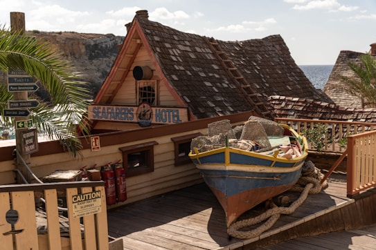 A rustic seaside scene featuring a wooden structure with signs pointing to different attractions such as restaurants and shops. A colorful old boat with fishing gear is positioned on the wooden deck, surrounded by various signs and palm trees. The backdrop includes shingled roofs and the ocean under a partly cloudy sky.