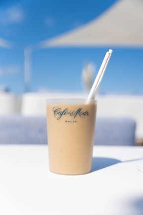 A beige drink in a transparent plastic cup with two white straws is placed on a white surface. The cup has the logo and text 'Café del Mar Malta' printed on it in blue. The background shows a sunny, clear blue sky and blurred white and blue elements, likely representing a coastal or outdoor café setting.