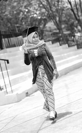 A person in graduation attire holding a cap. The scene is set outdoors with steps and trees in the background. The image is in black and white, capturing a sense of movement as the person walks forward.