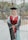 A person is standing outdoors wearing a graduation gown and cap, smiling confidently. The gown is primarily dark with light blue and maroon accents. The background shows a blurred view of trees and outdoor elements, suggesting a park or campus setting.