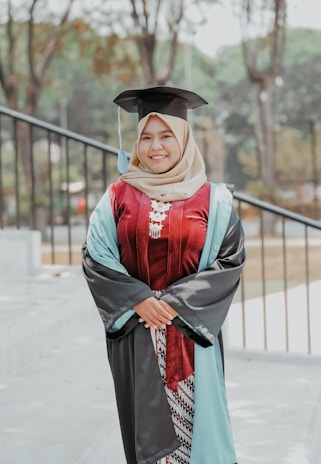 A person is standing outdoors wearing a graduation gown and cap, smiling confidently. The gown is primarily dark with light blue and maroon accents. The background shows a blurred view of trees and outdoor elements, suggesting a park or campus setting.