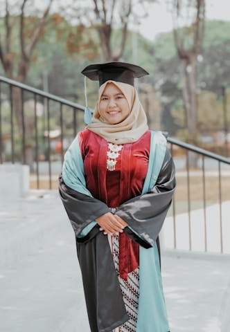 A person is standing outdoors wearing a graduation gown and cap, smiling confidently. The gown is primarily dark with light blue and maroon accents. The background shows a blurred view of trees and outdoor elements, suggesting a park or campus setting.