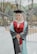 A person is standing outdoors wearing a graduation gown and cap, smiling confidently. The gown is primarily dark with light blue and maroon accents. The background shows a blurred view of trees and outdoor elements, suggesting a park or campus setting.