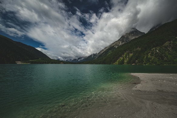 A serene lake stretches across the foreground, reflecting the deep green of the surrounding forest-covered mountains. The sky is dramatic, with a mix of dark and light clouds creating a striking contrast. The shoreline is visible with hints of sand or pebbles in the bottom right corner, suggesting a peaceful and untouched natural setting.