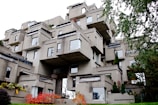 A unique, modern architectural structure composed of stacked concrete modules, creating an irregular, geometric appearance. The building features numerous windows and small terraces, with patches of greenery interspersed throughout the structure. Surrounding the building, foliage includes trees and shrubs with some displaying autumn colors.