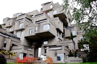 A unique, modern architectural structure composed of stacked concrete modules, creating an irregular, geometric appearance. The building features numerous windows and small terraces, with patches of greenery interspersed throughout the structure. Surrounding the building, foliage includes trees and shrubs with some displaying autumn colors.