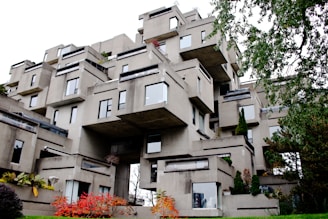A unique, modern architectural structure composed of stacked concrete modules, creating an irregular, geometric appearance. The building features numerous windows and small terraces, with patches of greenery interspersed throughout the structure. Surrounding the building, foliage includes trees and shrubs with some displaying autumn colors.