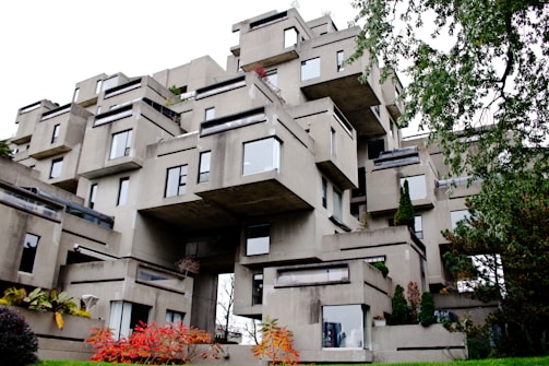 A unique, modern architectural structure composed of stacked concrete modules, creating an irregular, geometric appearance. The building features numerous windows and small terraces, with patches of greenery interspersed throughout the structure. Surrounding the building, foliage includes trees and shrubs with some displaying autumn colors.