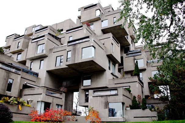 A unique, modern architectural structure composed of stacked concrete modules, creating an irregular, geometric appearance. The building features numerous windows and small terraces, with patches of greenery interspersed throughout the structure. Surrounding the building, foliage includes trees and shrubs with some displaying autumn colors.