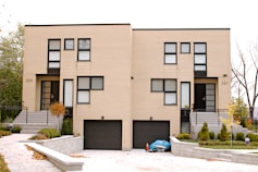 A modern, symmetrical duplex with beige brick walls and black-framed windows. Two identical front doors are visible, each accompanied by a small staircase. Below, there are two black garage doors. The landscaping features manicured bushes and small trees, with a paved driveway leading to the house.