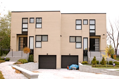 A modern, symmetrical duplex with beige brick walls and black-framed windows. Two identical front doors are visible, each accompanied by a small staircase. Below, there are two black garage doors. The landscaping features manicured bushes and small trees, with a paved driveway leading to the house.
