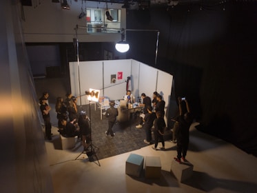 A group of people are gathered in a studio setting with lighting equipment and a backdrop. They appear to be engaged in a photo or video shoot, with several individuals holding cameras or adjusting lighting. The room is dimly lit, with a bright spotlight on the subject of the shoot. The backdrop is white, and there are softbox lights and other equipment visible.