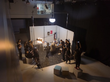 A group of people are gathered in a studio setting with lighting equipment and a backdrop. They appear to be engaged in a photo or video shoot, with several individuals holding cameras or adjusting lighting. The room is dimly lit, with a bright spotlight on the subject of the shoot. The backdrop is white, and there are softbox lights and other equipment visible.