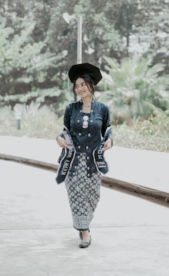 A person is wearing a traditional outfit and graduation cap, holding a sash. The backdrop consists of greenery and a paved pathway.