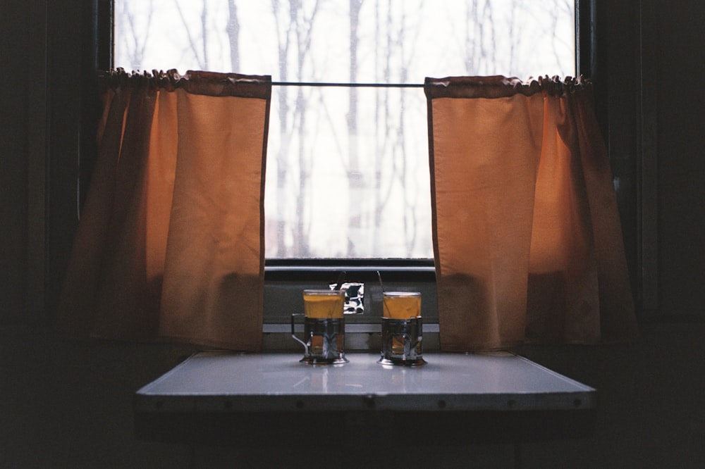 two glasses of orange juice sit on a table in front of a window