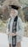 A person is wearing a graduation cap and a traditional outfit, holding a graduation sash with their name and academic achievement written on it. The background is lush and green, suggesting an outdoor location, possibly a garden or park.