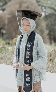 A person is wearing a graduation cap and a traditional outfit, holding a graduation sash with their name and academic achievement written on it. The background is lush and green, suggesting an outdoor location, possibly a garden or park.