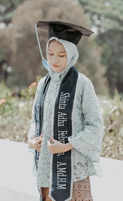 A person is wearing a graduation cap and a traditional outfit, holding a graduation sash with their name and academic achievement written on it. The background is lush and green, suggesting an outdoor location, possibly a garden or park.