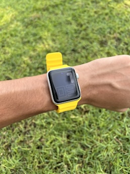 A smart watch with a yellow strap is worn on a wrist, set against a background of green grass.