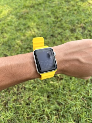 A smart watch with a yellow strap is worn on a wrist, set against a background of green grass.