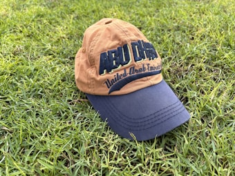 A baseball cap with the words 'Abu Dhabi' and 'United Arab Emirates' embroidered on it rests on a patch of lush green grass. The cap is two-toned, with a tan crown and a navy blue brim.
