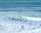 Surfers are scattered throughout a vibrant ocean with varied shades of blue and green. Waves create white foam as they crash towards the shore, while individuals with surfboards wade and position themselves in the water, indicating an active surfing scene.