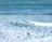 Surfers are scattered throughout a vibrant ocean with varied shades of blue and green. Waves create white foam as they crash towards the shore, while individuals with surfboards wade and position themselves in the water, indicating an active surfing scene.