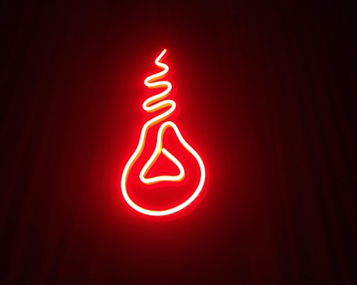 A bright red neon light is shaped in a unique design resembling a bulb with a twisted filament inside. It stands out vividly against a dark background, creating a striking visual effect.