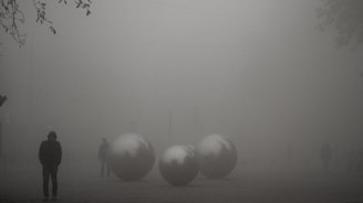 Foggy outdoor scene with large metallic spheres on the ground. Silhouetted figures walk in the background, creating a mysterious and surreal atmosphere. Sparse tree branches are visible in the upper left corner.