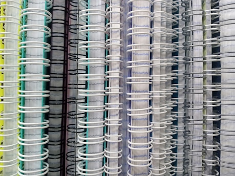 Several spiral-bound notebooks are arranged vertically side by side. The focus is on the spines, which are made of wire binding in various colors, including white, black, and gray. The covers of the notebooks display a range of colors such as yellow, green, and blue.