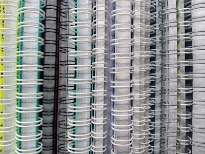 Several spiral-bound notebooks are arranged vertically side by side. The focus is on the spines, which are made of wire binding in various colors, including white, black, and gray. The covers of the notebooks display a range of colors such as yellow, green, and blue.