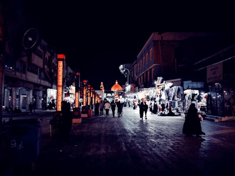 A bustling night market scene with illuminated storefronts and vibrant lights. People are casually walking down a wide, paved street lined with shops on either side. The architecture includes various building styles, and the focus draws towards a distant, ornately lit dome.
