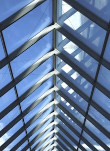 A series of triangular glass panels form a modern, symmetric roof structure. The panels allow natural light to stream through, and the clear blue sky is visible beyond. Metal framing holds the glass panes in place, creating a clean and geometric pattern accented by cables.