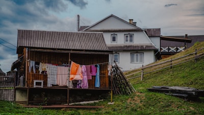 A rustic rural scene featuring a wooden house with a corrugated iron roof and a larger white house in the background. Clothes are hanging on a line to dry, and a wooden fence runs along a grassy slope. The sky is cloudy, suggesting overcast weather.