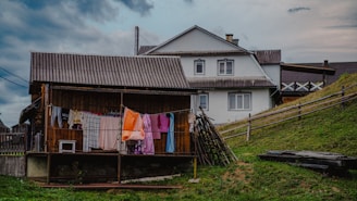 A rustic rural scene featuring a wooden house with a corrugated iron roof and a larger white house in the background. Clothes are hanging on a line to dry, and a wooden fence runs along a grassy slope. The sky is cloudy, suggesting overcast weather.