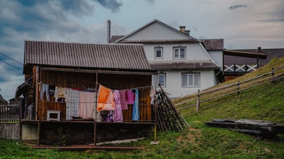 A rustic rural scene featuring a wooden house with a corrugated iron roof and a larger white house in the background. Clothes are hanging on a line to dry, and a wooden fence runs along a grassy slope. The sky is cloudy, suggesting overcast weather.