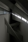 Concrete beams intersect at various angles within a minimalist, industrial space. Natural light filters through a row of windows, casting geometric shadows on the walls.