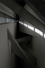 Concrete beams intersect at various angles within a minimalist, industrial space. Natural light filters through a row of windows, casting geometric shadows on the walls.