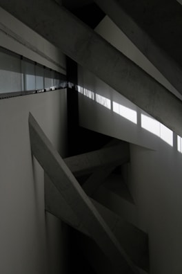 Concrete beams intersect at various angles within a minimalist, industrial space. Natural light filters through a row of windows, casting geometric shadows on the walls.